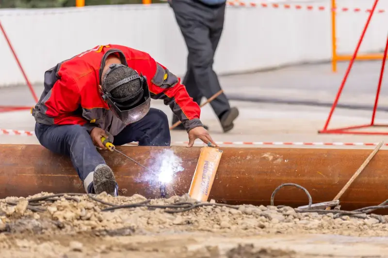 Welder in a mask kneeling on a steel pipe at an outdoor site