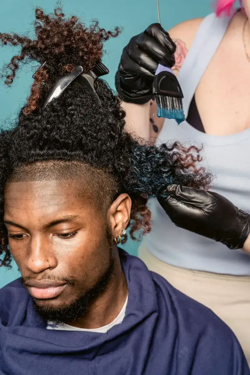 Stylist in black gloves painting blue color into a man's curly hair