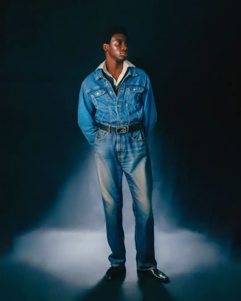 Male model in a light-wash denim jacket and wide jeans standing in a spotlight