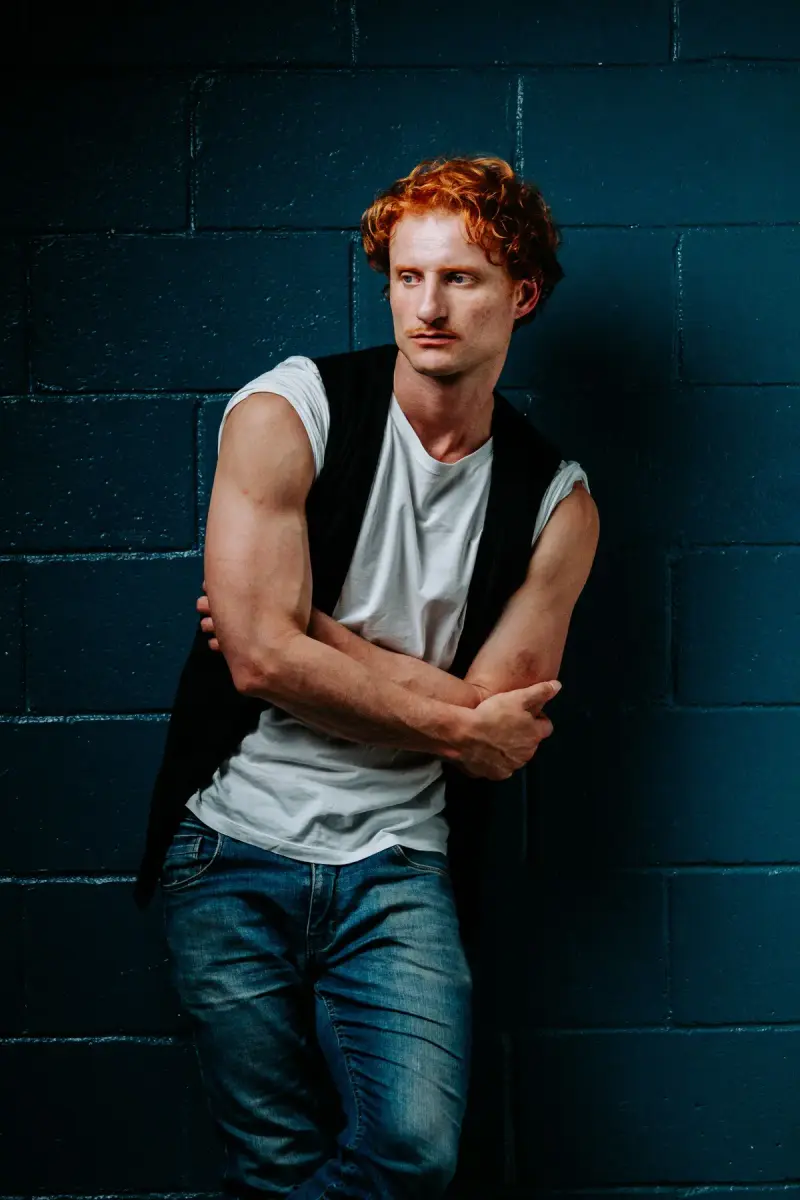Red-haired male model in a black vest, gray tee and dark jeans against a blue wall