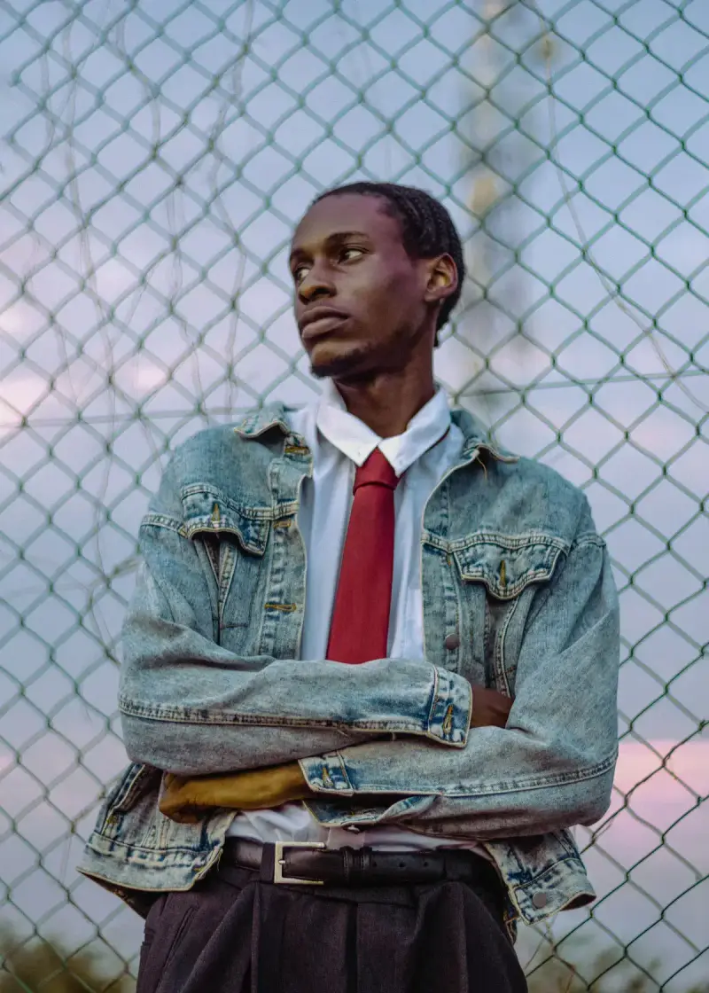 Man in a faded denim jacket, white shirt and red tie by a chain-link fence at dusk