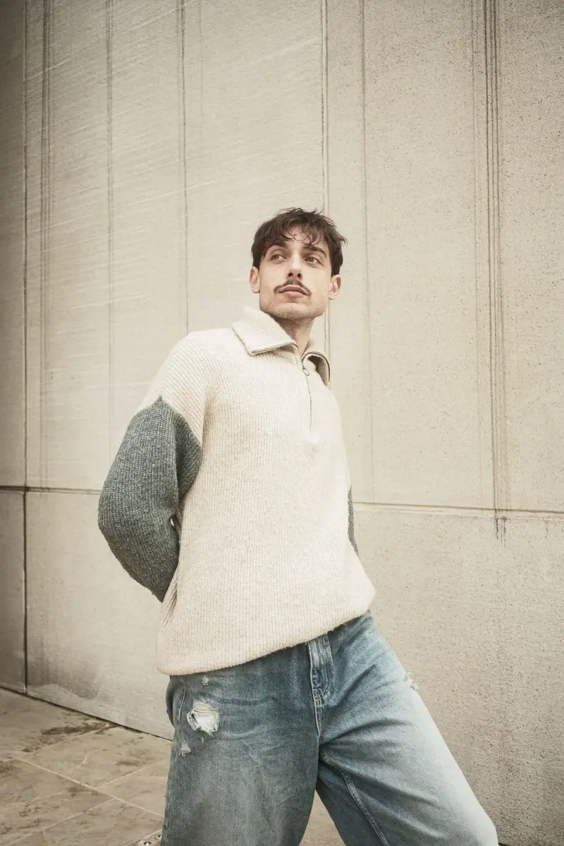 Young man in a cream color-block sweater and ripped jeans leaning on a concrete wall