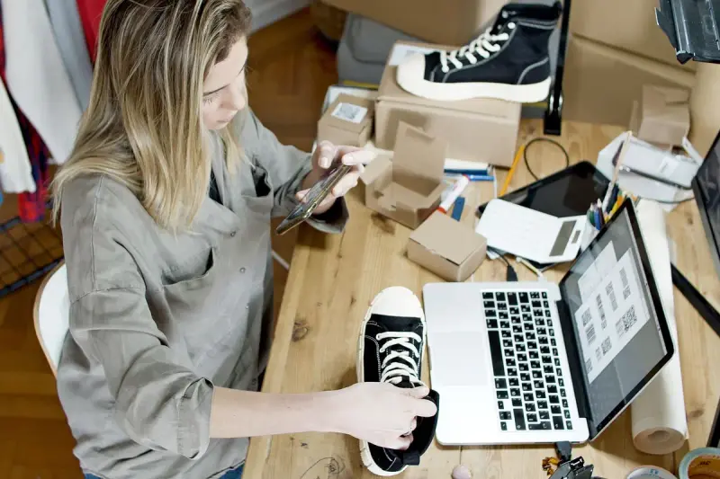 Seller photographing a sneaker on her desk beside a laptop and packing materials