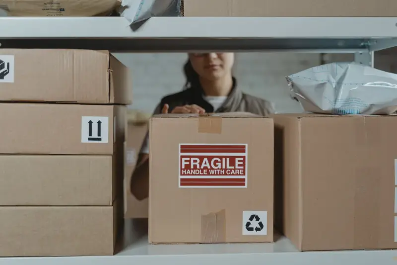 Cardboard boxes stacked on a shelf, one marked fragile, with a worker packing behind
