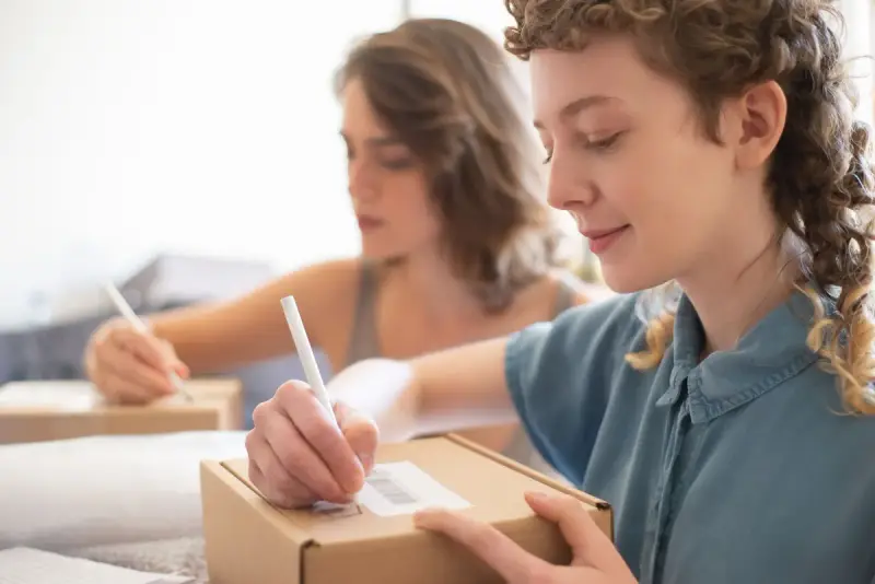 Two young women writing shipping details on a cardboard box