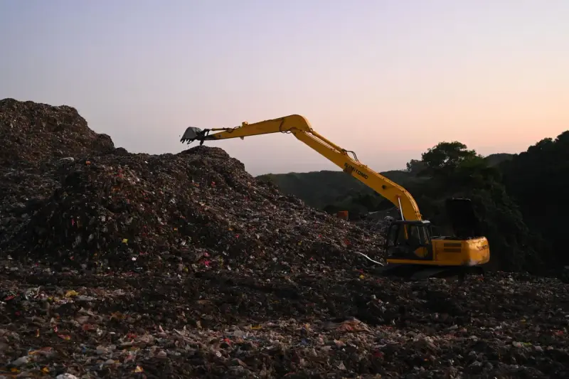 Yellow long-reach excavator on a landfill mound at dusk