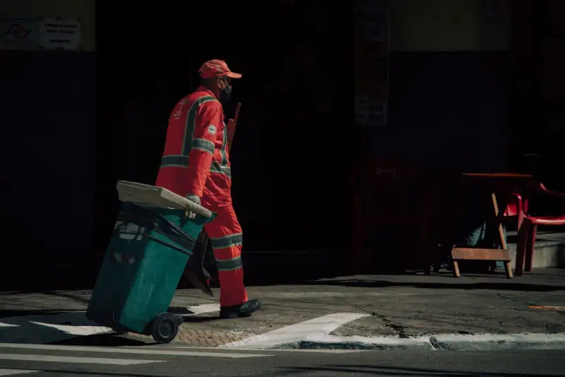 Sanitation worker in red coveralls wheeling a green waste cart along a street