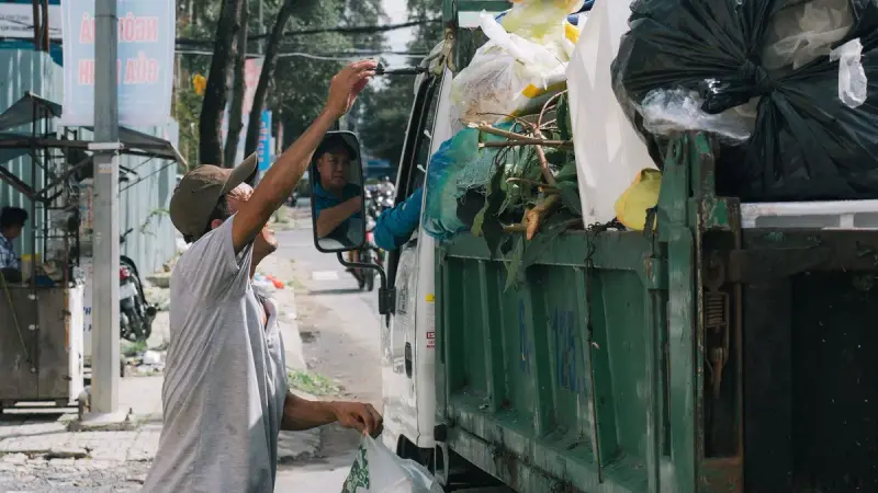 Worker in a cap lifting a bag of trash into the back of a collection truck