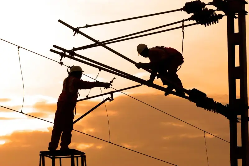 Two line workers silhouetted against a sunset sky while repairing power lines