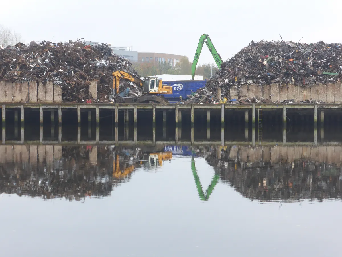 Waterfront scrap metal yard with two large piles, a material handler and an excavator reflected in still water