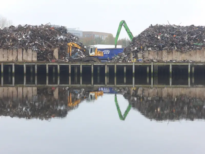 Waterfront scrap metal yard with two large piles, a material handler and an excavator reflected in still water