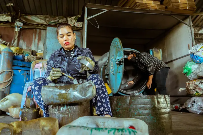 Woman in work gloves cutting a used plastic container while a coworker loads a machine behind her