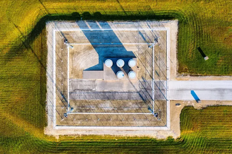 Aerial view of a fenced gas facility with small white tanks in a green field
