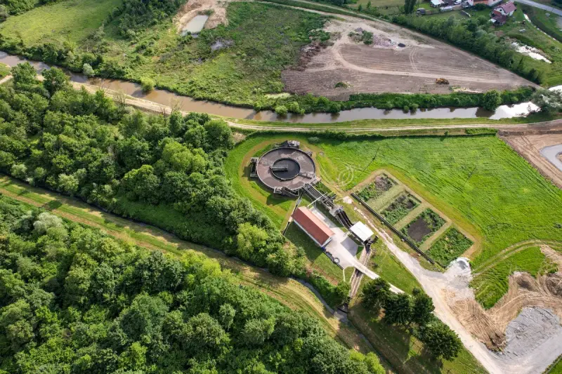 Aerial view of a small wastewater treatment plant with round tanks beside a river and fields