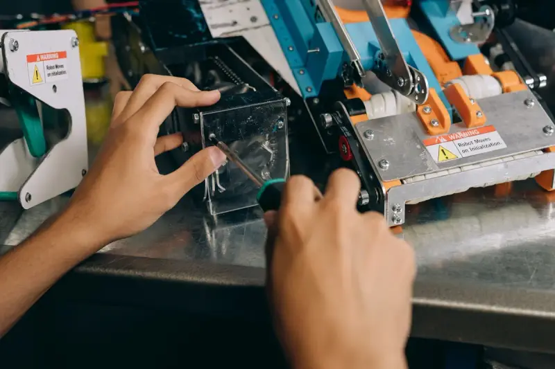 Hands adjusting a clear guard on a robot mechanism with warning labels on a steel bench