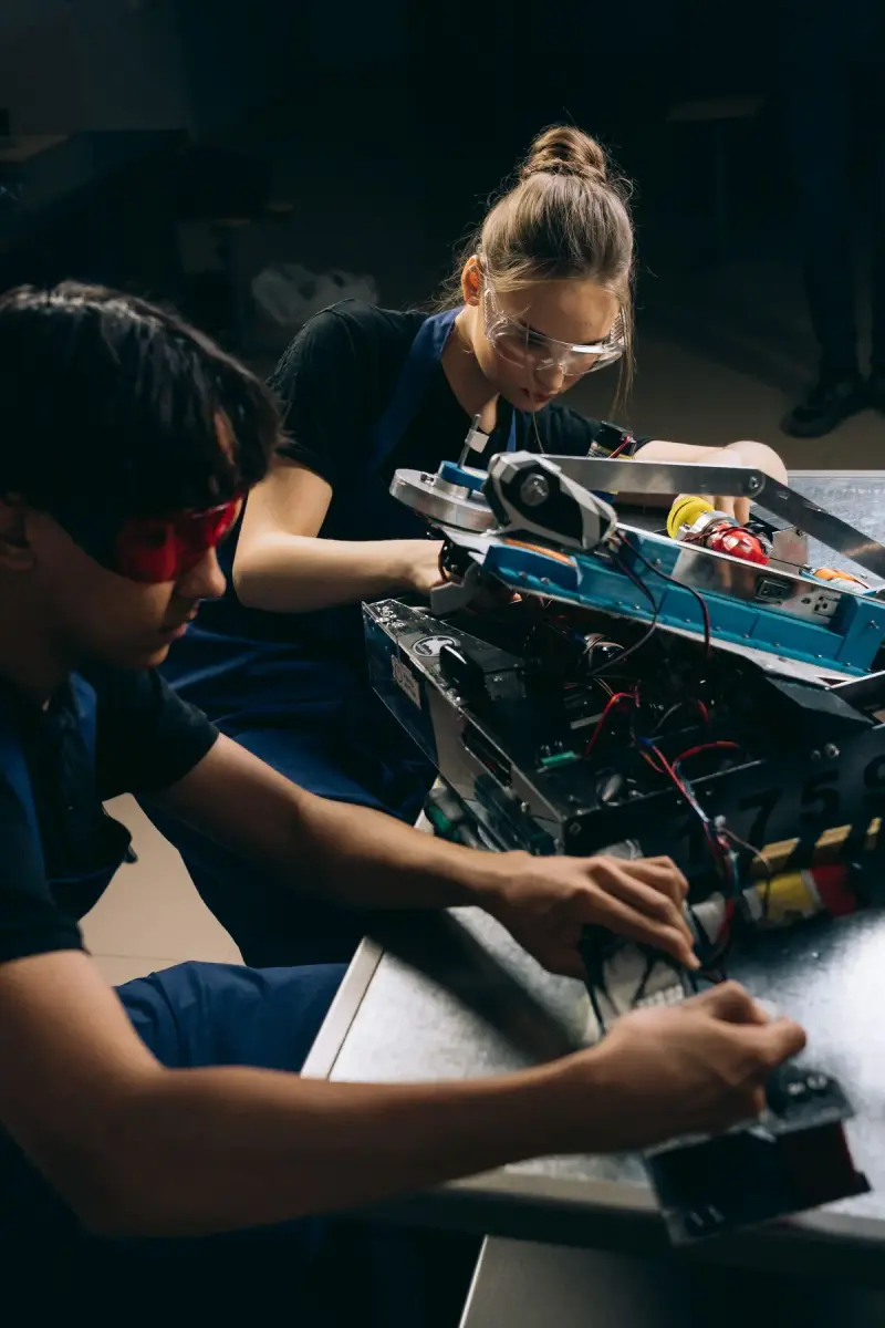 Young woman and man in safety glasses wiring and assembling a robot chassis at a workbench