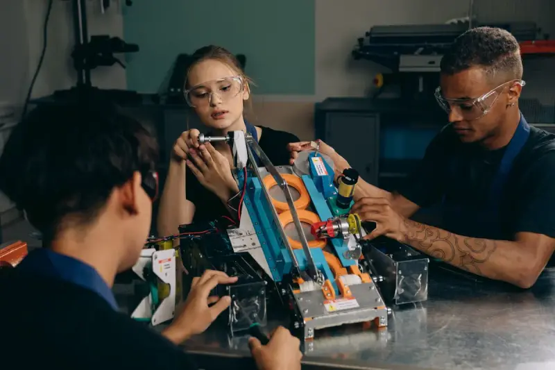 Three young engineers in safety glasses assembling a colorful robot on a workshop bench