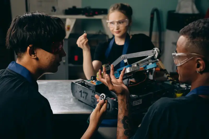 Three young engineers in safety glasses assembling a robot with an arm mechanism at a workbench