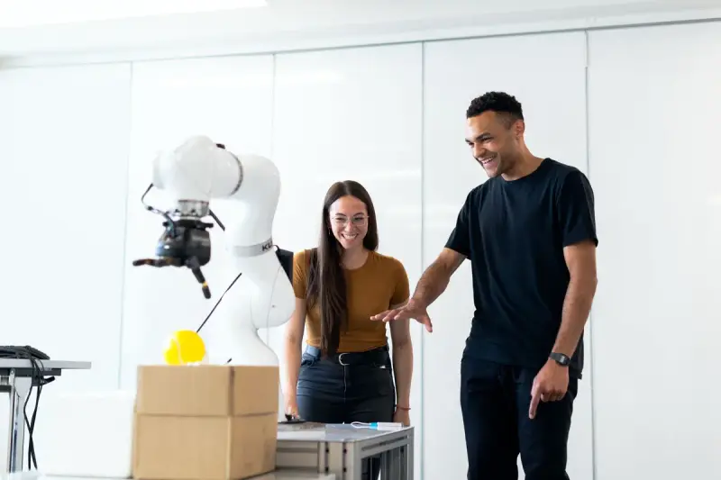Two smiling engineers watching a white robot arm lift a part in a bright lab