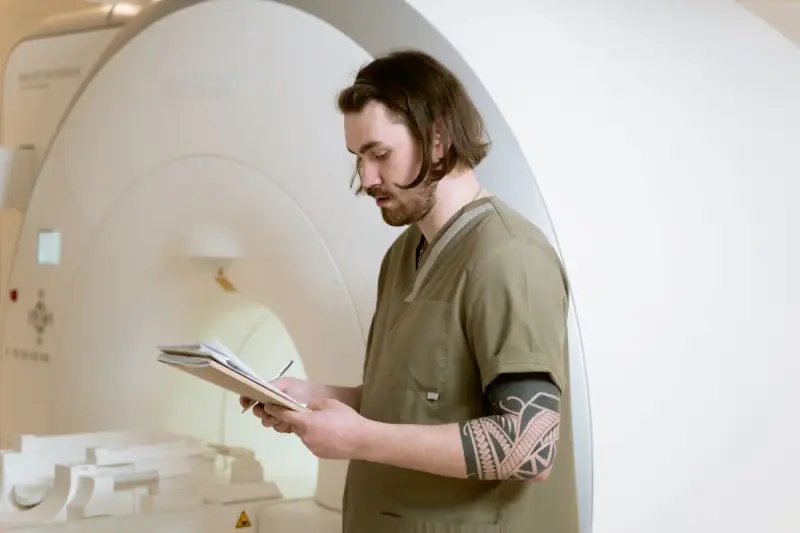Radiologic technologist in scrubs taking notes beside an MRI scanner