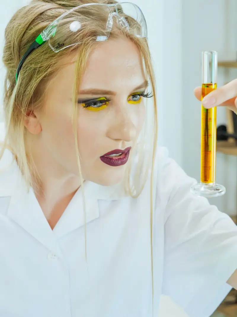 Woman with yellow eyeliner and safety glasses examining an amber graduated cylinder