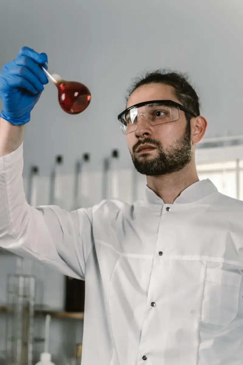 Bearded chemist in safety glasses and lab coat holding a flask of red liquid