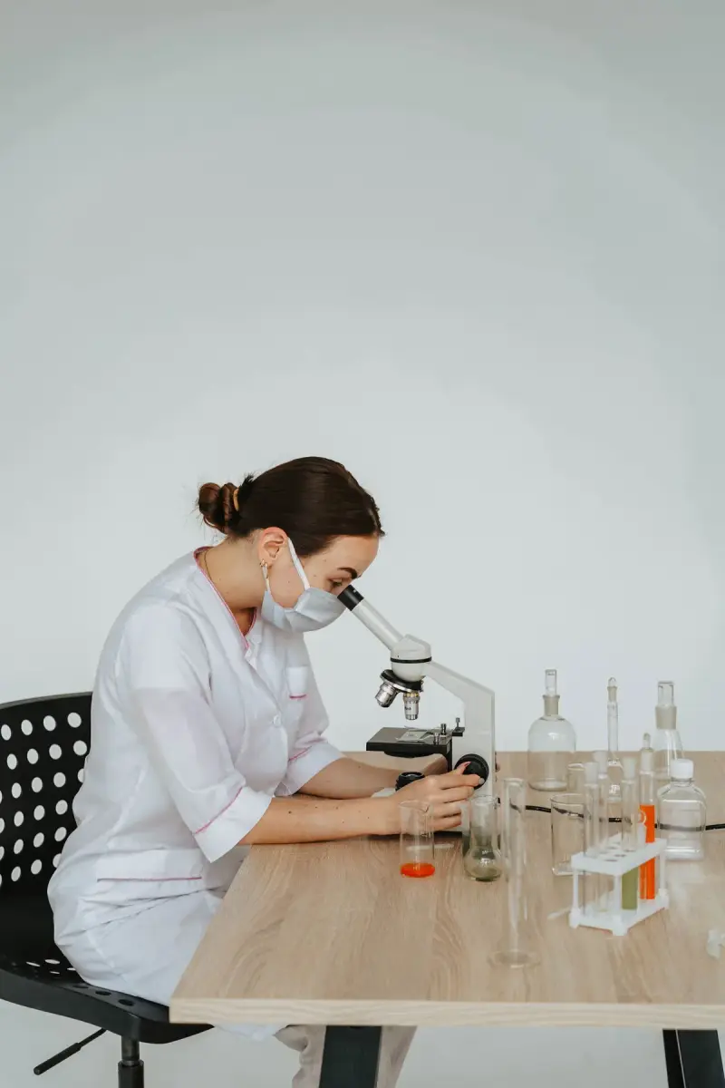Scientist in a white coat and mask looking through a microscope
