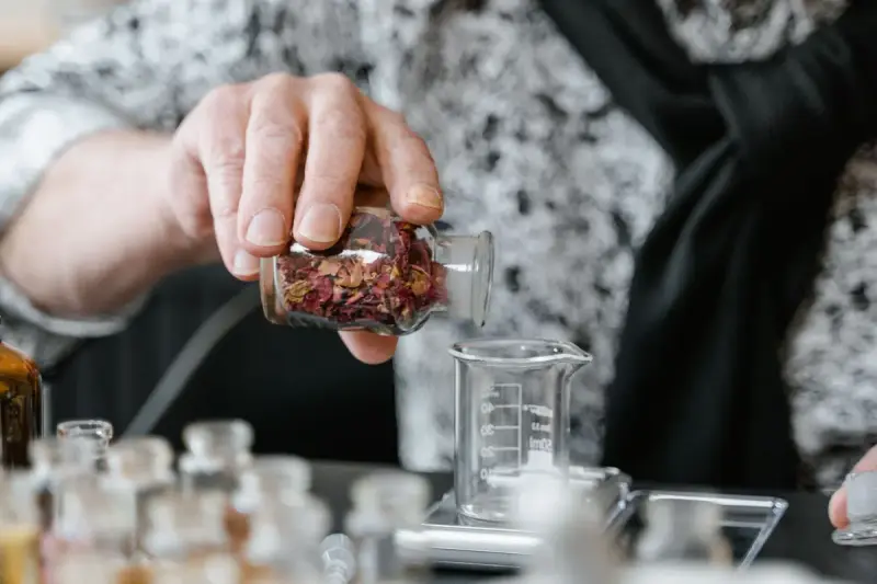 Hand pouring dried botanicals from a glass bottle beside lab beakers