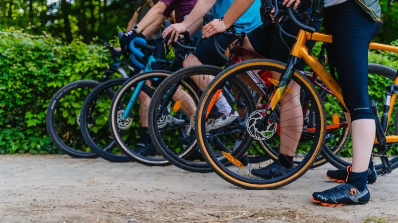 Row of riders standing with gravel bikes on a dirt path beside green hedges