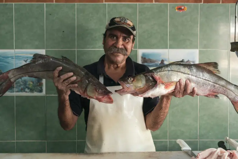 Smiling fishmonger in a white apron holding up two large fresh fish in his shop