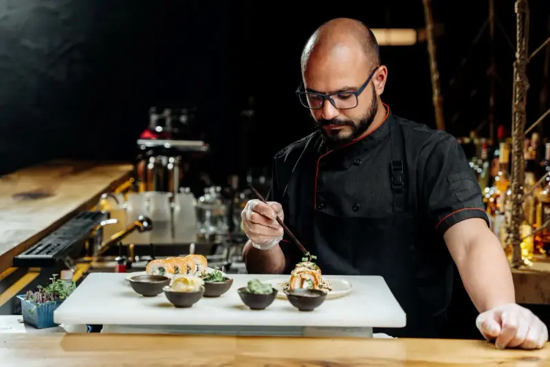 Chef in a black jacket and gloves plating sushi rolls with tweezers in a restaurant