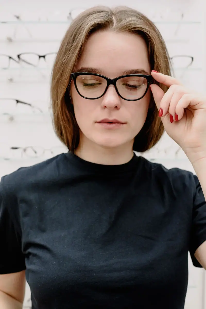Woman adjusting black cat-eye eyeglasses in front of an optical store frame wall