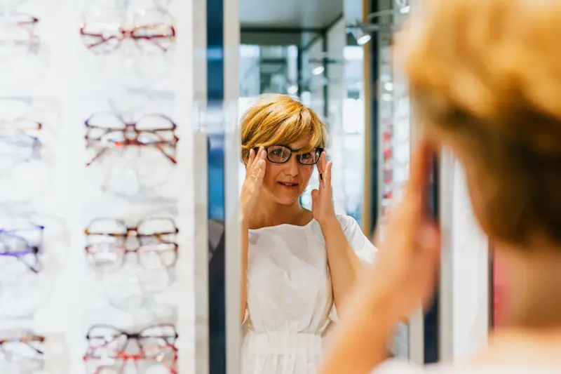 Woman trying on black eyeglasses in a mirror beside an optical frame wall
