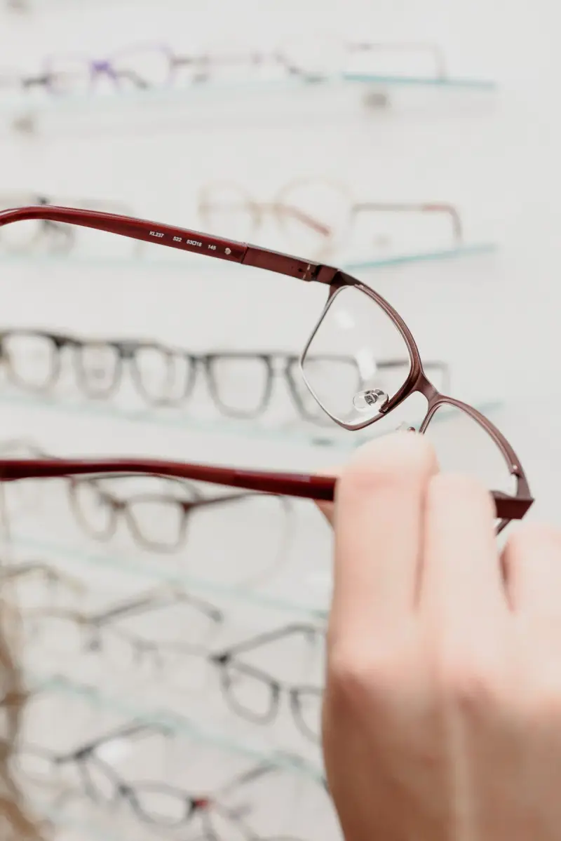 Hand lifting burgundy metal eyeglass frames from a glass display shelf