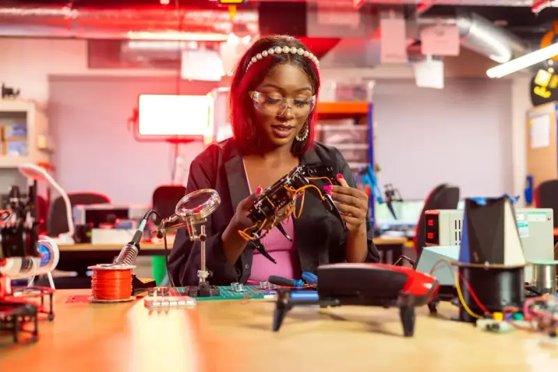 A woman works with a small drone and electronic components at a workbench.