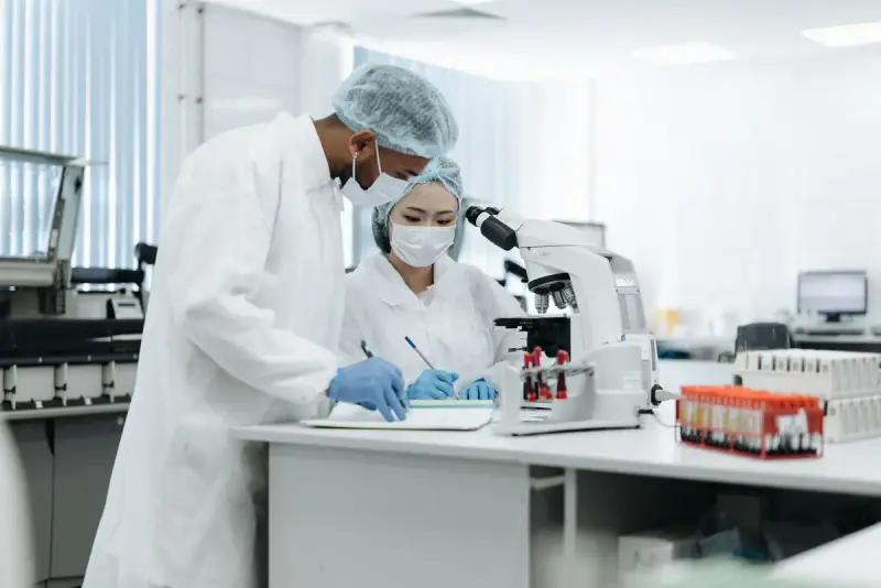 Two researchers in lab coats and hair nets working at a microscope bench in a bright lab