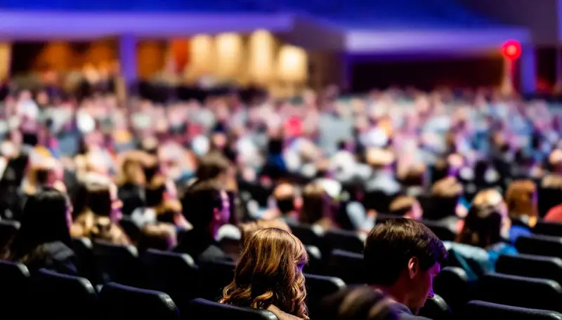 Large crowd seated in a conference hall listening to a session under colored lights