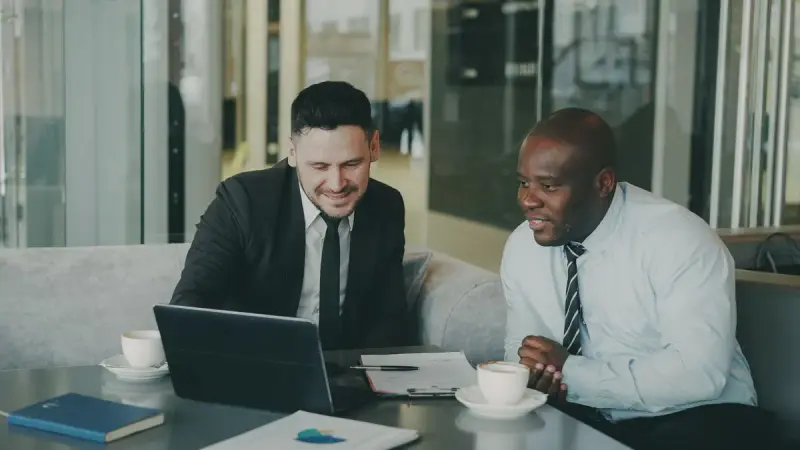 Two businessmen smiling over a laptop and coffee in a modern office lounge
