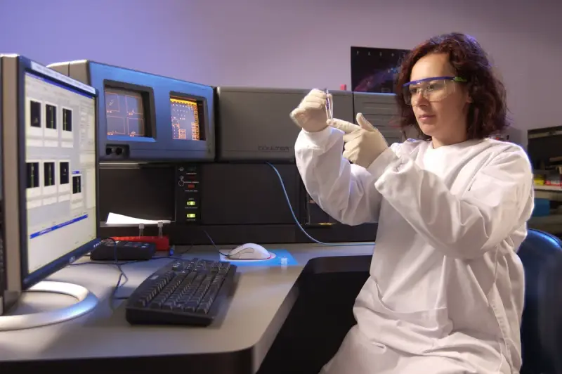 Scientist in a lab coat and safety glasses preparing a sample tube at a flow cytometer workstation