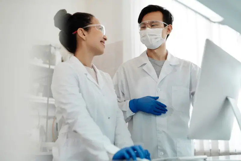 Smiling female scientist and masked male colleague in lab coats talking beside a monitor