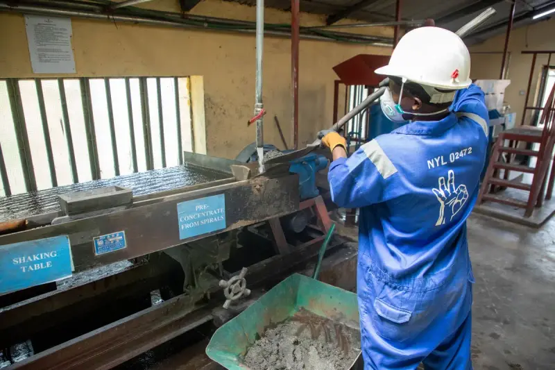 Worker in blue coveralls, hard hat and mask running a shaking table that separates ore concentrate