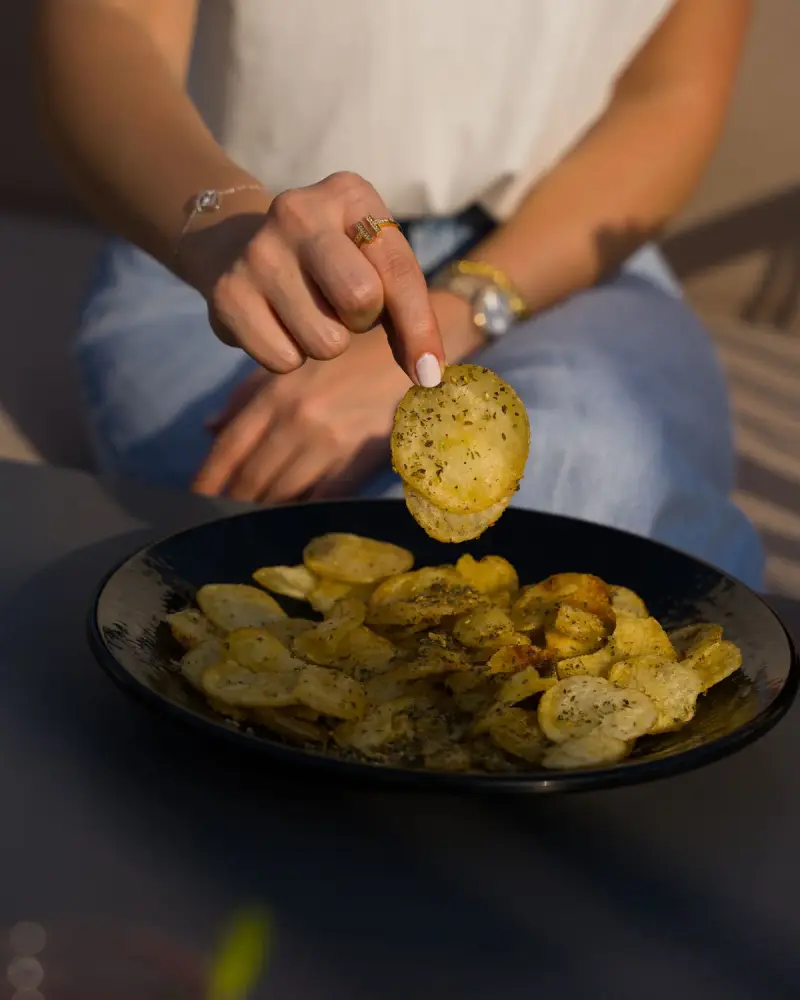 Hand lifting a herb-seasoned potato chip above a dark bowl full of chips