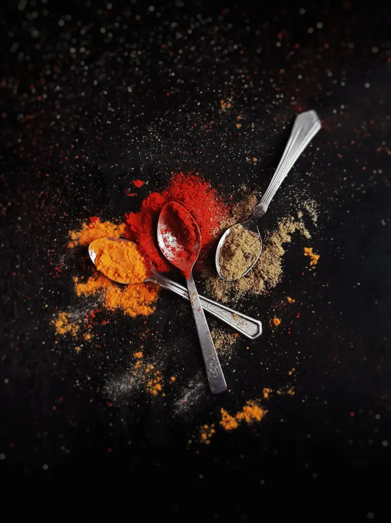 Three metal spoons holding red, orange and brown seasoning powders on a dark surface