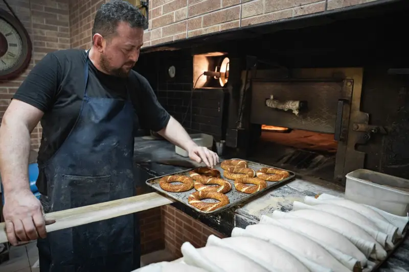 Baker in a dark apron slides a tray of sesame-crusted bread rings into a brick oven