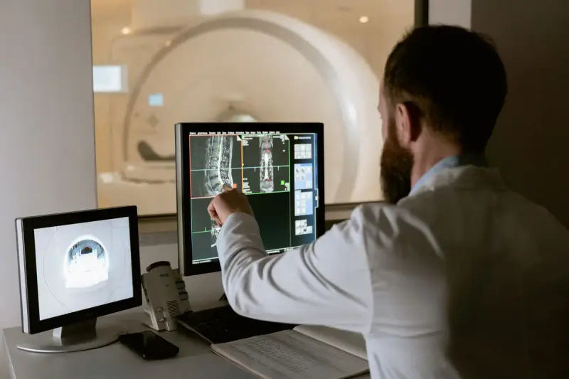 Clinician in a white coat pointing at scan images on a monitor, with a scanner behind the control-room window