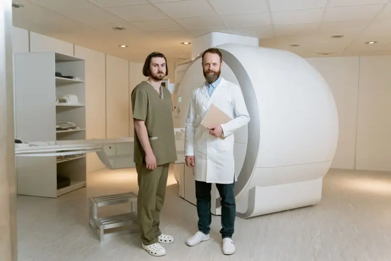 Technologist in scrubs and physician in a white coat standing beside a large imaging scanner