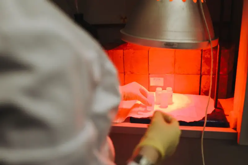 Gloved hands handling small sample vials under a lamp in front of stacked bricks