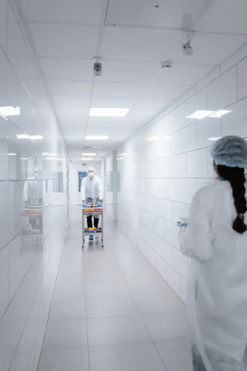 Woman in a cleanroom gown and cap watching a colleague push a cart of sample tubes down a white corridor