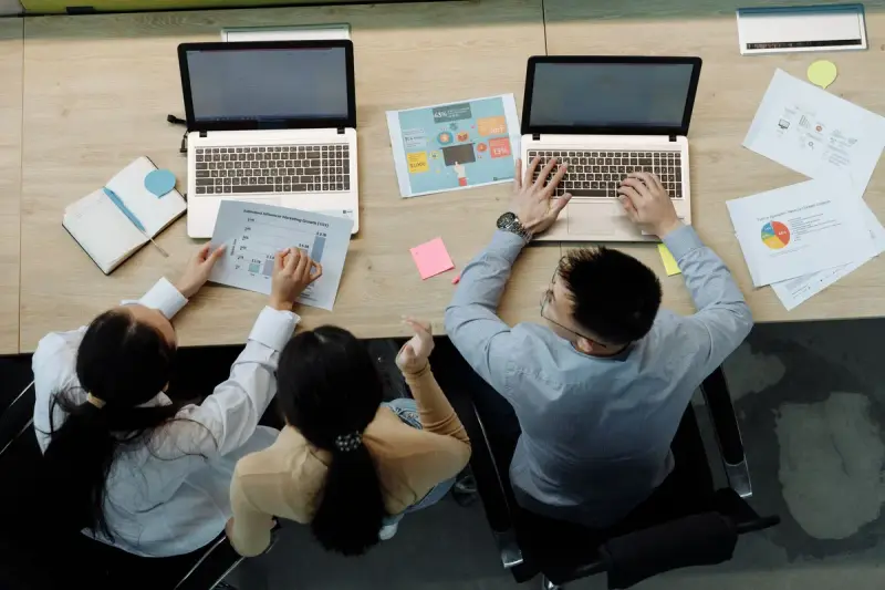Three colleagues working on laptops and documents at a shared desk