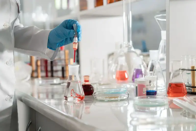 Gloved hand using a dropper above flasks of colored liquids on a lab bench